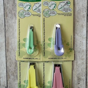 May include: Four Clover fusible bias tape makers in green, purple, yellow, and pink. Each tool is packaged on a card with instructions and the Clover logo. The tools are designed to create fusible bias tapes.