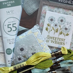 May include: A collection of cross-stitch supplies, including fabric, embroidery floss, and a framed piece with the text "Sweetest of Wishes." The floss colors range from yellow-green to gray. The fabric is marked with Japanese text.