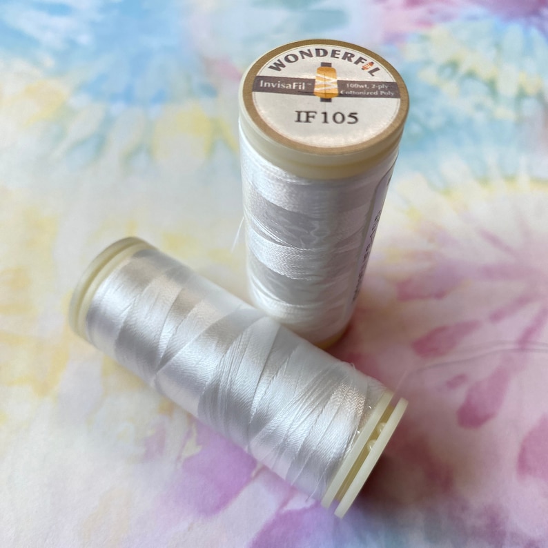 Op de afbeelding: Twee spoelen witte draad, met de labels "Invisafill" en "IF 105", met de woorden "Wonderful 100wt, 2 ply, Cotonized Poly" op het label.