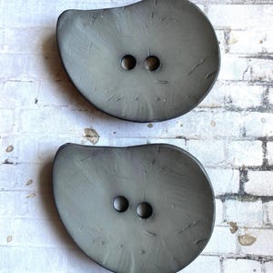 May include: Two large, irregular, organic-shaped buttons in a muted, semi-translucent gray tone. Each button features a smooth, slightly textured surface with subtle, darker gray inclusions and two centered holes for attachment.