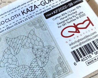 Puede incluir: Un primer plano de una tela blanca con un diseño de bordado sashiko negro. La tela está etiquetada como "Sashiko Cloth "Kaza-Guruma 5"  ITEM #KF2022-38 GREIGE". La etiqueta también incluye información sobre la composición de la tela, el diseñador y el fabricante.