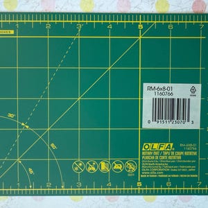 May include: A green OLFA rotary mat, 8 inches by 6 inches, with yellow grid lines and measurement markings. The mat includes angled lines for precise cutting and a label with product information and a barcode.