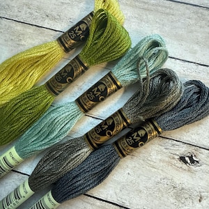 May include: A collection of six skeins of DMC embroidery floss in various shades of green, blue, and gray. Each skein is labeled with "MC 25" and "8.7 yd". The floss is arranged on a light wood grain surface.