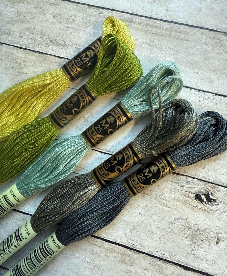 May include: A collection of six skeins of DMC embroidery floss in various shades of green, blue, and gray. Each skein is labeled with "MC 25" and "8.7 yd". The floss is arranged on a light wood grain surface.