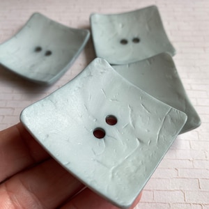 May include: Four square, light gray buttons with two holes each. The buttons have a textured surface and a slightly rounded shape. The buttons are a pale, muted gray color.