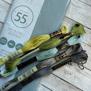May include: A package of light blue cross-stitch fabric with the text "55" and "14 ct" printed on the packaging. Several skeins of embroidery floss in shades of green, blue, and gray are arranged next to the fabric.