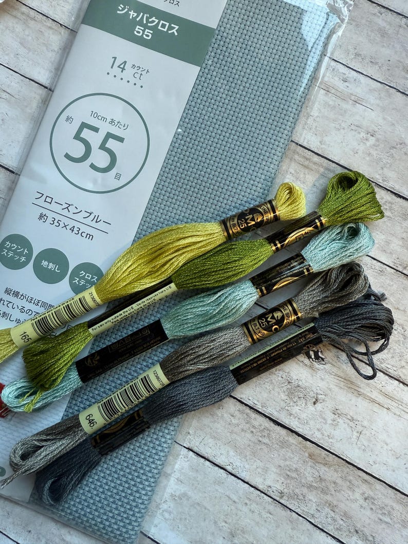 May include: A package of light blue cross-stitch fabric with the text "55" and "14 ct" printed on the packaging. Several skeins of embroidery floss in shades of green, blue, and gray are arranged next to the fabric.