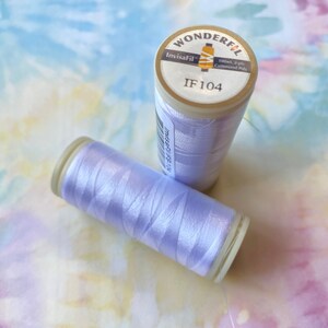 Op de afbeelding: Een spoel lichtpaarse draad met het label "InvisaFil IF 104", het merk "WonderFil" en de tekst "100wt 2-ply Cottonized Poly".