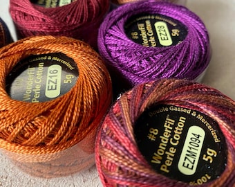 May include: Close-up of several spools of WonderFil Perle Cotton thread in various colors, including orange, purple, and burgundy. Each spool is labeled with the thread type, weight, and color code. The thread is tightly wound and appears smooth.