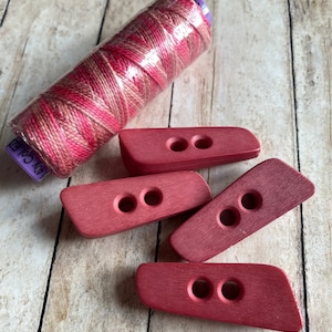 May include: Four red wooden toggle buttons with two holes each. A spool of pink and brown embroidery floss is in the background. The floss label reads "100% Cotton".
