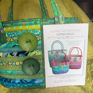 May include: A quilted fabric bag in shades of green, blue, and yellow with two large green buttons on the front. Beside the bag is a pattern instruction sheet titled Aunties Two Patterns Camden Bags, showing images of three different colored bags.