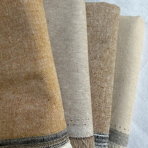 K&ouml;nnte beinhalten: Vier gefaltete rechteckige Stoffst&uuml;cke in den Farben Senfgelb, Hellbeige, Hellbraun und Cremewei&szlig;. Jedes St&uuml;ck weist eine gewebte Textur auf und ist am unteren Rand mit einem dunklen horizontalen Streifen sowie kurzen, ausgefransten wei&szlig;en Fransen versehen.