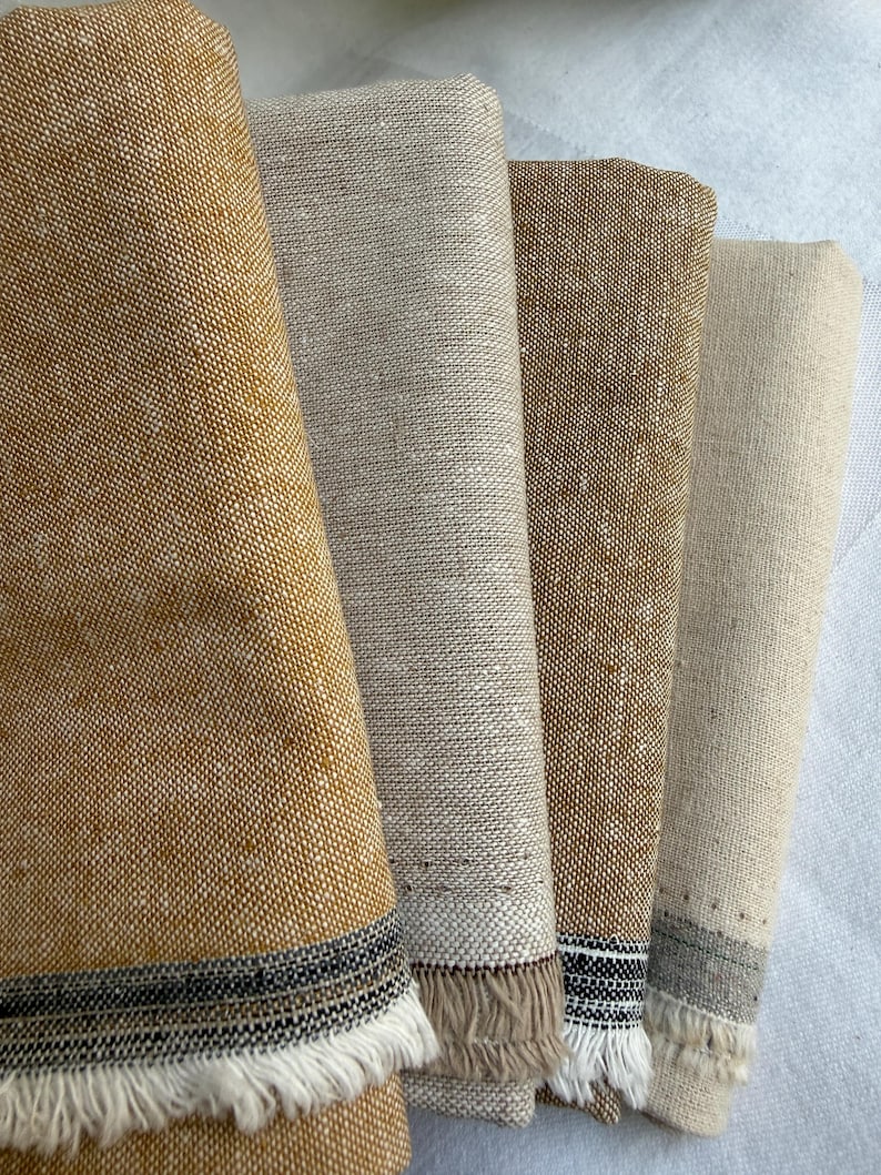 K&ouml;nnte beinhalten: Vier gefaltete rechteckige Stoffst&uuml;cke in den Farben Senfgelb, Hellbeige, Hellbraun und Cremewei&szlig;. Jedes St&uuml;ck weist eine gewebte Textur auf und ist am unteren Rand mit einem dunklen horizontalen Streifen sowie kurzen, ausgefransten wei&szlig;en Fransen versehen.