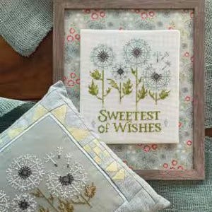 May include: A framed cross-stitch featuring dandelions with the text "Sweetest of Wishes." A matching pillow with a dandelion design is in the foreground. The image also includes the text "Dandelion Wishes" and the Robin Pickens Cross Stitch logo.