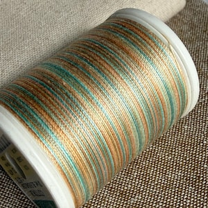 May include: A spool of variegated sewing thread featuring a repeating color palette of soft tan, warm copper, and muted teal. The thread is wound tightly around a white plastic cylinder, displaying a consistent striped pattern across the entire surface.