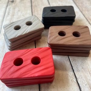 May include: Four stacks of two-hole rectangular buttons in red, brown, beige, and black. The buttons have a textured surface and are stacked on top of each other.