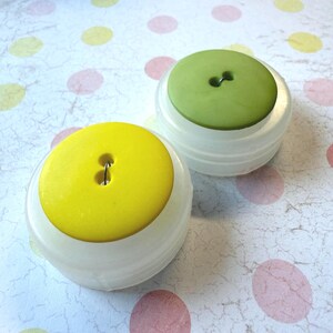 May include: Two decorative button-shaped objects. One has a bright yellow top, the other a light green top. Both sit atop white, rounded bases. The background is a patterned paper with pink and yellow polka dots.