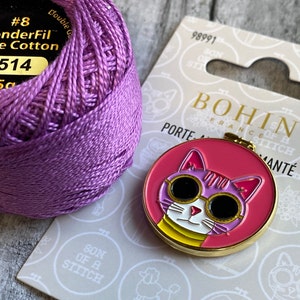 Pode incluir: Um carretel de linha de algod&atilde;o roxa ao lado de um bastidor de bordado redondo rosa e dourado em forma de gato. O gato usa &oacute;culos de sol e o texto "BOHIN FRANCE".