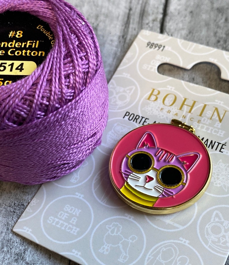 Pode incluir: Um carretel de linha de algod&atilde;o roxa ao lado de um bastidor de bordado redondo rosa e dourado em forma de gato. O gato usa &oacute;culos de sol e o texto "BOHIN FRANCE".