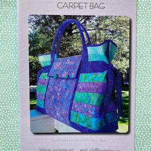 May include: A sewing pattern for a carpet bag. The pattern is printed on a light pink background with a picture of a purple and green carpet bag with a flap and a button closure. The text on the pattern reads "Aunties Two Patterns Carpet Bag" and "Copyright 2014 Pattern Number AT 292".