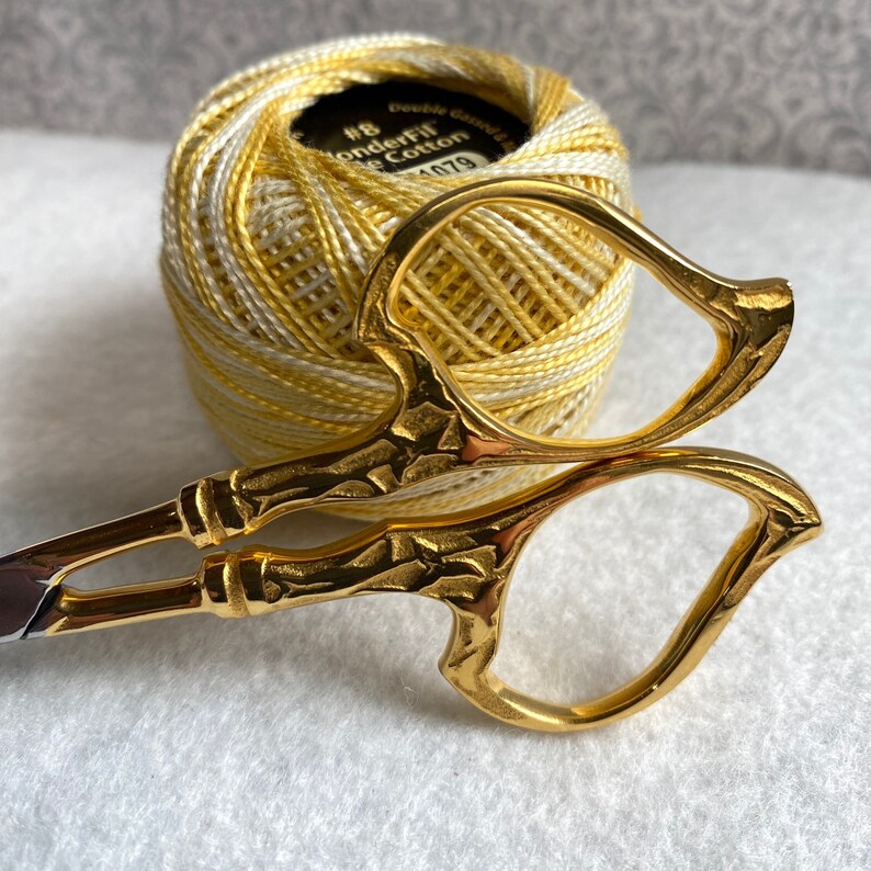 Pode incluir: Uma tesoura de bordar dourada com uma al&ccedil;a decorativa repousa em frente a uma bola de fio de algod&atilde;o WonderFil amarelo e branco. A bola de fio tem uma etiqueta preta com texto branco. A tesoura tem um design detalhado e texturizado.