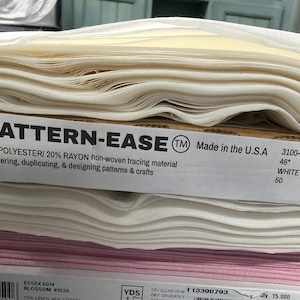 May include: A stack of folded fabric bolts including white Pattern-Ease non-woven tracing material composed of 80% polyester and 20% rayon, measuring 46 inches wide, positioned above a bolt of pink fabric with a visible product label.