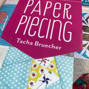 May include: A pink book titled "Handy Pocket Guide Paper Piecing" by Tacha Brueche. The book cover features a patchwork design with a variety of colors and patterns.