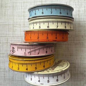 May include: A stack of six spools of ribbon with a ruler printed on them. The spools are in various colors, including blue, white, orange, pink, yellow, and white. The ribbon is a woven, textured fabric.
