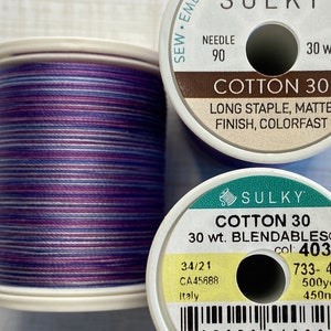 May include: Two spools of Sulky Cotton 30 thread. The spool on the left is a purple, white, and pink variegated thread. The spool on the right is a solid white spool with black text that reads "Sulky Cotton 30 30 wt. Blendables® col: 4032 733-4032 500yds 450m 34/21 CA45688 Italy".