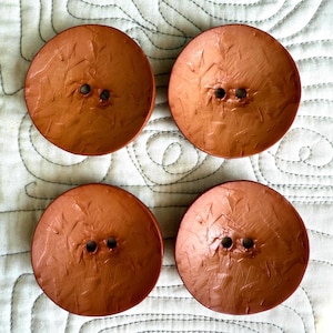 May include: Four round, terracotta-colored buttons arranged in a square formation. Each button features a slightly concave, textured surface with visible brushstroke-like markings and two centered holes for attachment to fabric or garments.
