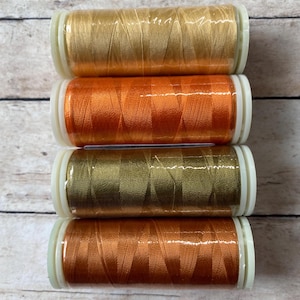 May include: Four spools of thread in shades of orange, gold, and brown. The spools are arranged vertically, with the gold spool on top and the brown spool on the bottom.