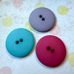 May include: Three large, round buttons in shades of teal, lavender, and magenta. Each button has two holes and a matte finish. The buttons are arranged on a patterned surface with pastel-colored polka dots.