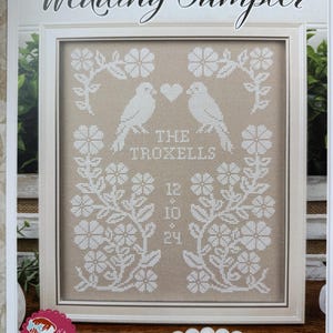 May include: A framed cross-stitch sampler with the text "Wedding Sampler." The design features two white birds facing each other above a heart, with floral and leaf accents. The text "THE TROXELLS" and a date are also included.