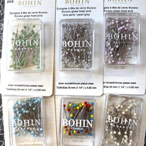 May include: Six clear plastic cases containing Bohin Murano glass head pins in green, pearl grey, lavender, light blue, multicolored, and cream. Each case is mounted on a white card labeled with nickel plated steel and a size of 30 mm (1 1/4 inches).