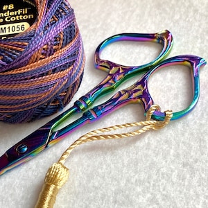May include: A pair of iridescent rainbow-colored embroidery scissors with a gold tassel attached. The scissors have a decorative design and are closed. A ball of purple, brown, and blue embroidery floss is in the background.