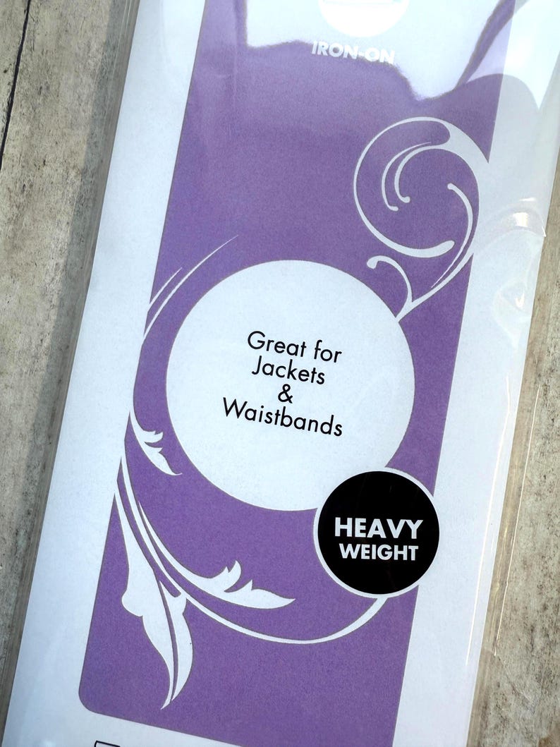 Op de afbeelding: Een rechthoekige verpakking van zware opstrijkbare tussenvoering met een paarse achtergrond en witte wervelende afbeeldingen. De tekst luidt: IRON-ON, Great for Jackets & Waistbands en HEAVY WEIGHT. De verpakking is van doorzichtig plastic.