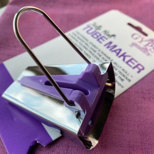 May include: A silver and purple tube maker tool with a curved metal handle. The tool has a purple plastic guide and is packaged with a white and purple card that reads "Tube Maker". The tool is designed for quilting.