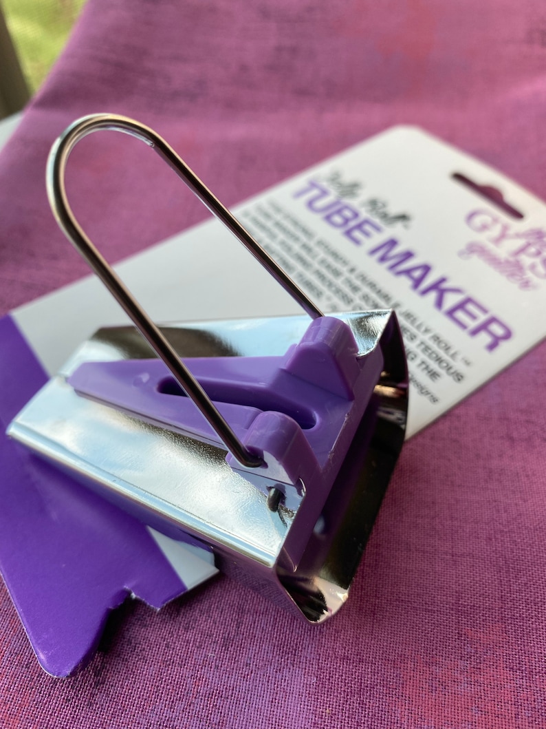 May include: A silver and purple tube maker tool with a curved metal handle. The tool has a purple plastic guide and is packaged with a white and purple card that reads "Tube Maker". The tool is designed for quilting.
