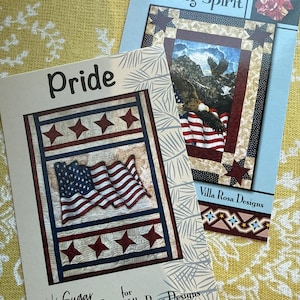May include: Two quilting pattern cards with a patriotic theme. The top card features a bald eagle flying over an American flag with the text "Daring Spirit" and "Villa Rosa Designs". The bottom card features a folded American flag with stars and the text "Pride" and "Sugar Pine Quilt Designs for Villa Rosa Designs".