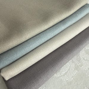 May include: A stack of four folded fabric swatches featuring a visible woven texture. The colors include a light beige, a soft pale blue, a neutral tan, and a dark charcoal gray, all layered neatly on top of each other in a diagonal arrangement.