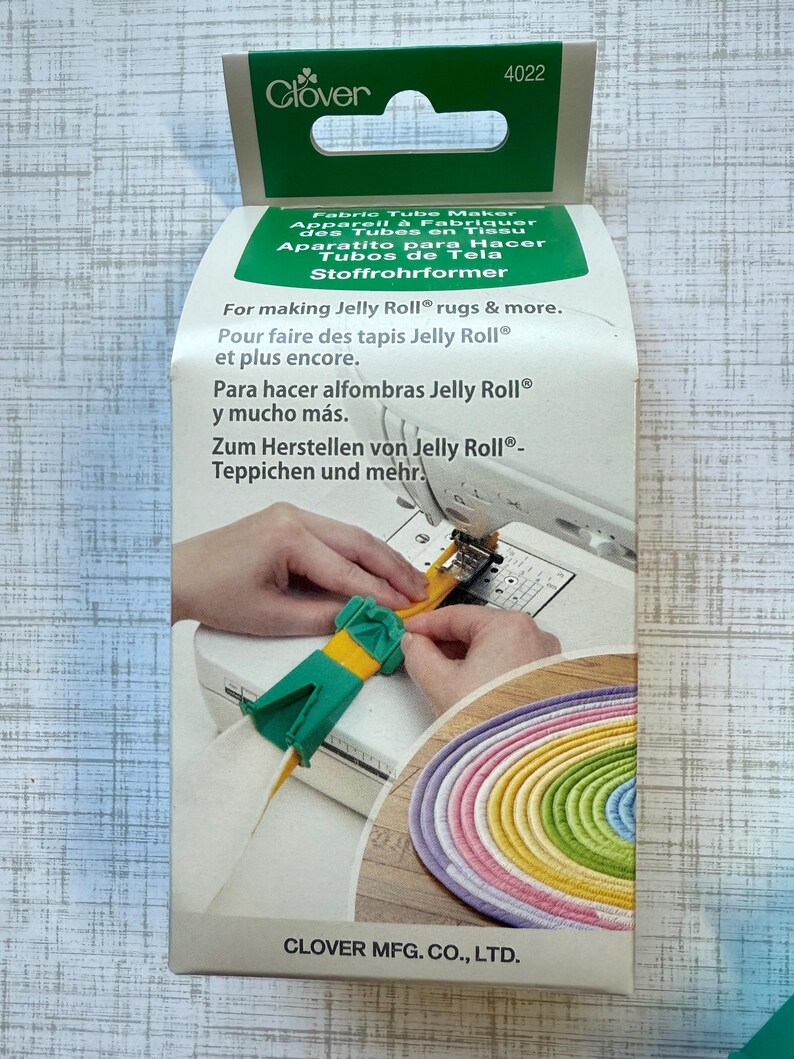 May include: A Clover brand fabric tube maker, model 4022, in a white and green cardboard box. The packaging features a yellow and green plastic tool being used with a sewing machine to create a coiled, multi-colored fabric rug.