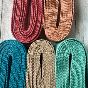 May include: Close-up of six spools of flat, woven ribbon in various colors. The colors include coral, peach, teal, tan, and seafoam green. The ribbons are arranged in a curved, U-shape, and are made of a textured material.