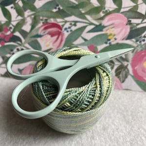 Pode incluir: Uma bola de linha verde e amarela com um par de pequenas tesouras verde-claro em cima. A linha &eacute; enrolada firmemente e as tesouras t&ecirc;m cabos arredondados e l&acirc;minas afiadas. O fundo apresenta um padr&atilde;o floral.