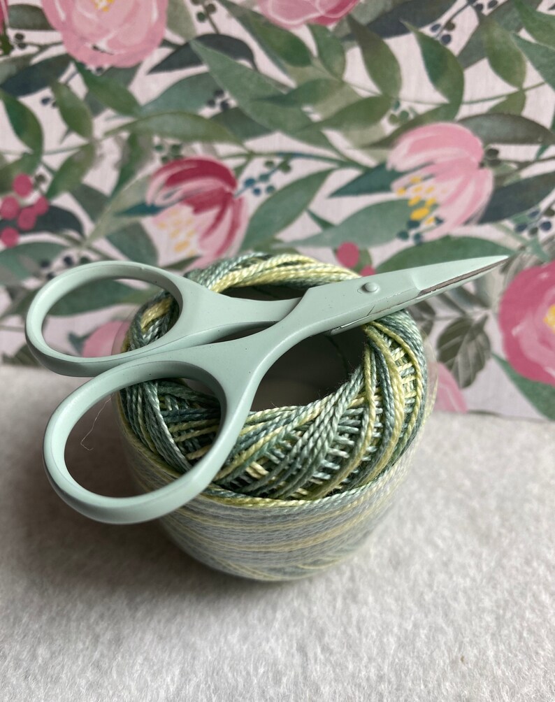Pode incluir: Uma bola de linha verde e amarela com um par de pequenas tesouras verde-claro em cima. A linha &eacute; enrolada firmemente e as tesouras t&ecirc;m cabos arredondados e l&acirc;minas afiadas. O fundo apresenta um padr&atilde;o floral.