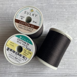 May include: Two spools of black Sulky Cotton 30 thread. The spool on the left is labeled "Sulky Cotton 30 Solid, col: 1234, 733-1234, 500yds, 450m". The spool on the right is labeled "Sulky Cotton 30, 30 wt., Long Staple, Matte Finish, Colorfast".