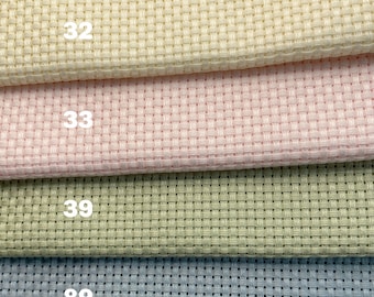 May include: Four different colors of Aida cloth, a type of fabric used for cross-stitch, are stacked on top of each other. The colors are cream, pink, green, and blue. The numbers 32, 33, 39, and 89 are printed on the fabric.