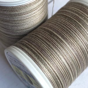 May include: Two spools of beige and brown thread with a subtle shimmer. The thread is wound around white plastic spools.