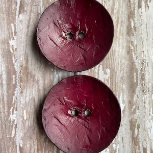 May include: Two large, round, burgundy buttons with two holes each. The buttons have a textured surface and are set against a weathered, light-colored wooden background. These buttons are ideal for sewing and craft projects.