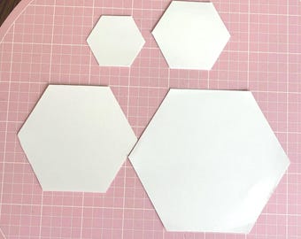 May include: Four white, flat, hexagonal shapes of varying sizes arranged on a pink grid surface. The shapes are solid white with smooth edges and a matte finish, featuring two smaller hexagons at the top and two larger hexagons positioned below them.
