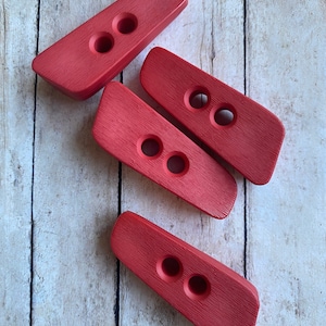 May include: Four red wooden toggle buttons with two holes each. The buttons are arranged in a random pattern on a white wooden surface.